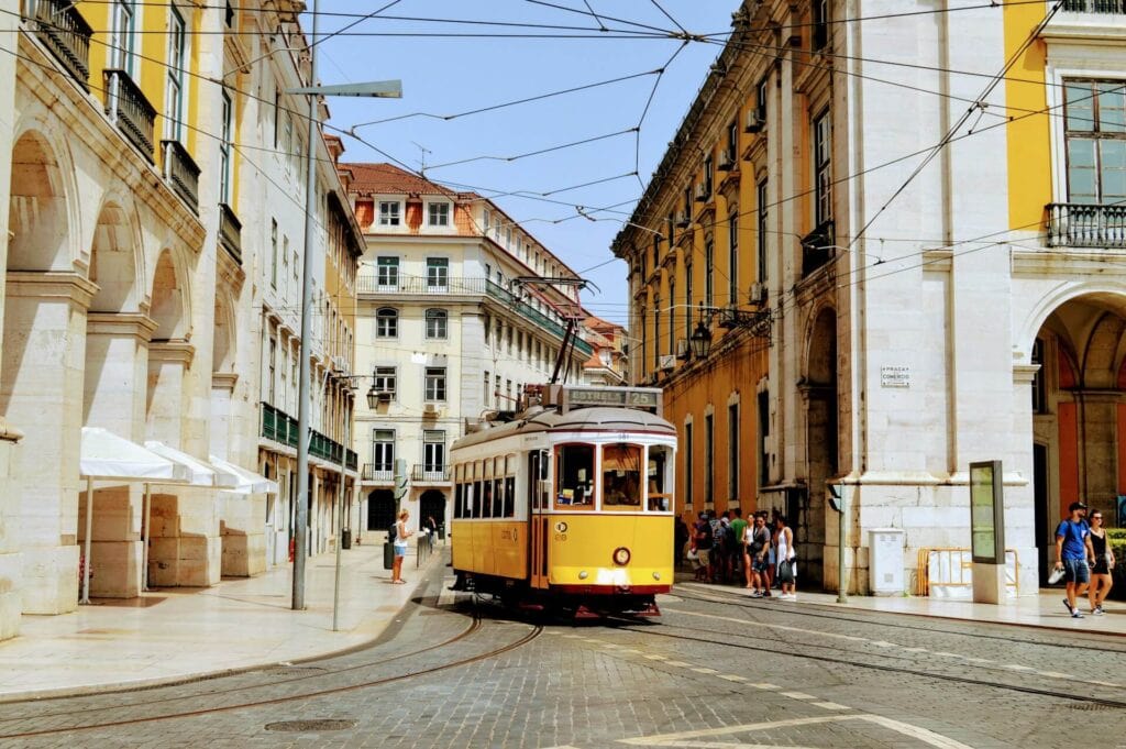 Eine historische gelbe Straßenbahn fährt durch eine belebte, von hellen klassizistischen Gebäuden gesäumte Straße mit Arkadengängen, in Lissabon, Portugal.