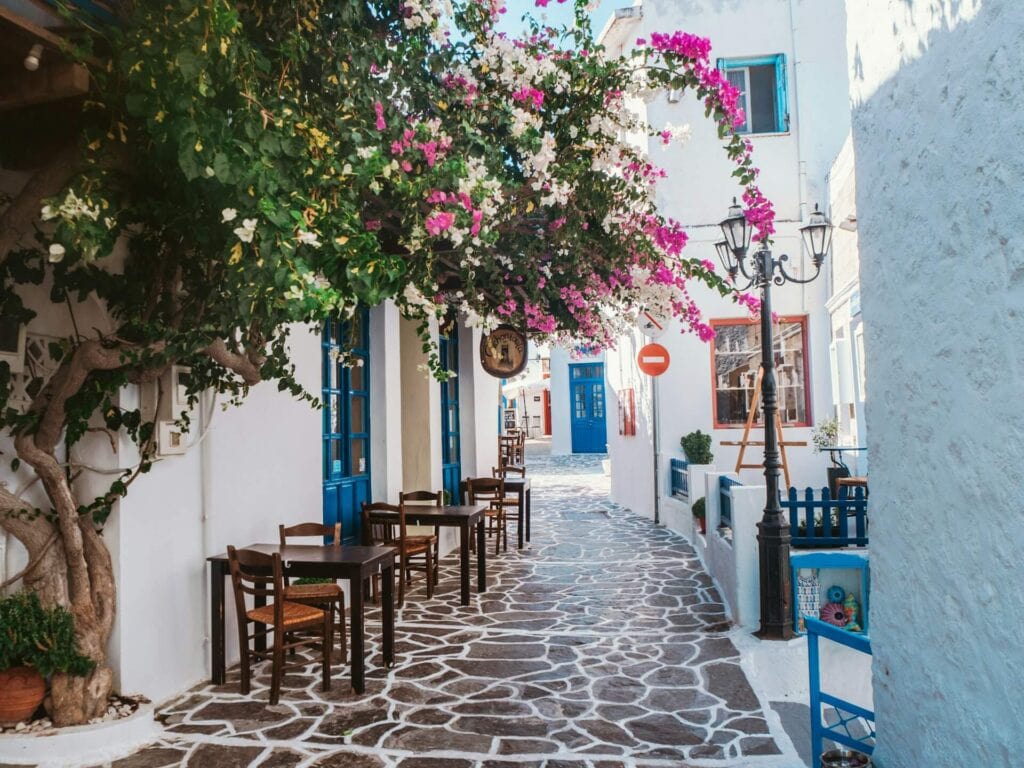 Eine schmale, malerische Gasse auf der griechischen Insel Milos. Weiße Häuser mit blauen Türen und Fenstern säumen den Weg aus unregelmäßigem Kopfsteinpflaster. Über den Tischen eines kleinen Cafés im Freien blühen üppige, pinkfarbene Bougainvillea-Blumen.