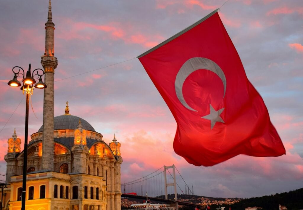 Die Ortaköy-Moschee in Istanbul bei einem rötlich-violetten Sonnenuntergang. Im Vordergrund weht eine große rote Nationalflagge der Türkei. Im Hintergrund sind die beleuchtete Bosporus-Brücke und das gegenüberliegende Ufer zu sehen