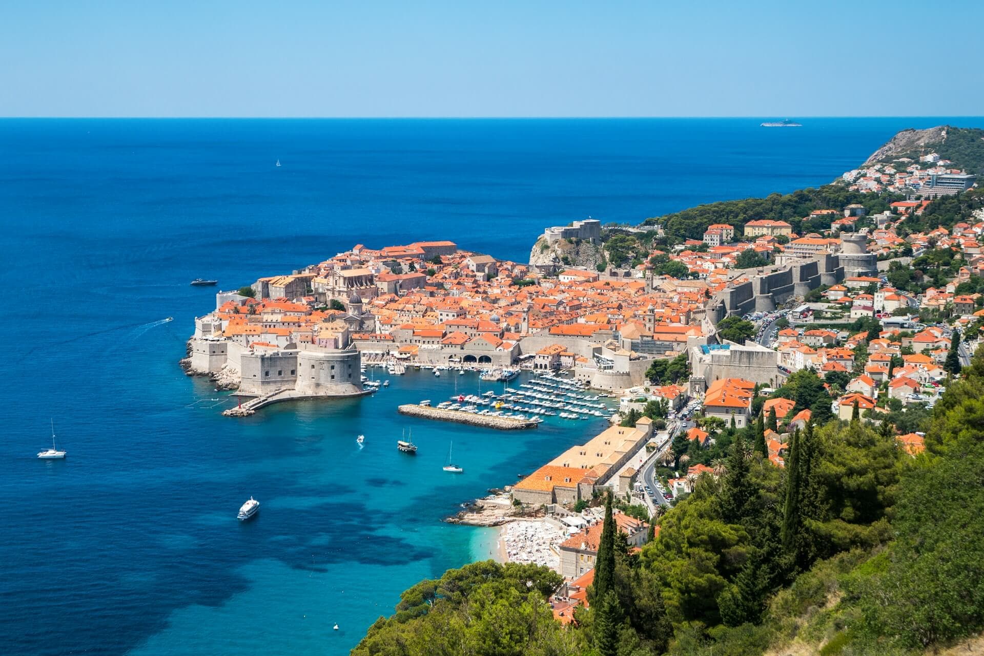 Panorama-Luftaufnahme der historischen Altstadt von Dubrovnik an der adriatischen Küste. Die mittelalterlichen Stadtmauern und Gebäude mit charakteristischen orangefarbenen Ziegeldächern ragen in das tiefblaue Meer hinein. Ein kleiner Hafen mit Yachten ist im Vordergrund sichtbar.