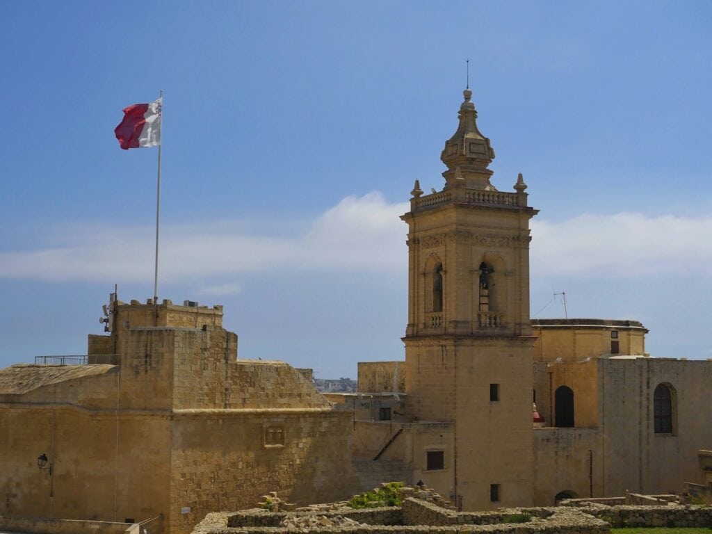 Blick auf die historische Cittadella in Victoria auf der maltesischen Insel Gozo. Eine große maltesische Flagge weht über den honigfarbenen Festungsmauern, daneben erhebt sich ein barocker Glockenturm aus Kalkstein. Im Vordergrund sind die alten Befestigungsanlagen zu sehen, darüber ein weiter Himmel mit dichten Wolken.