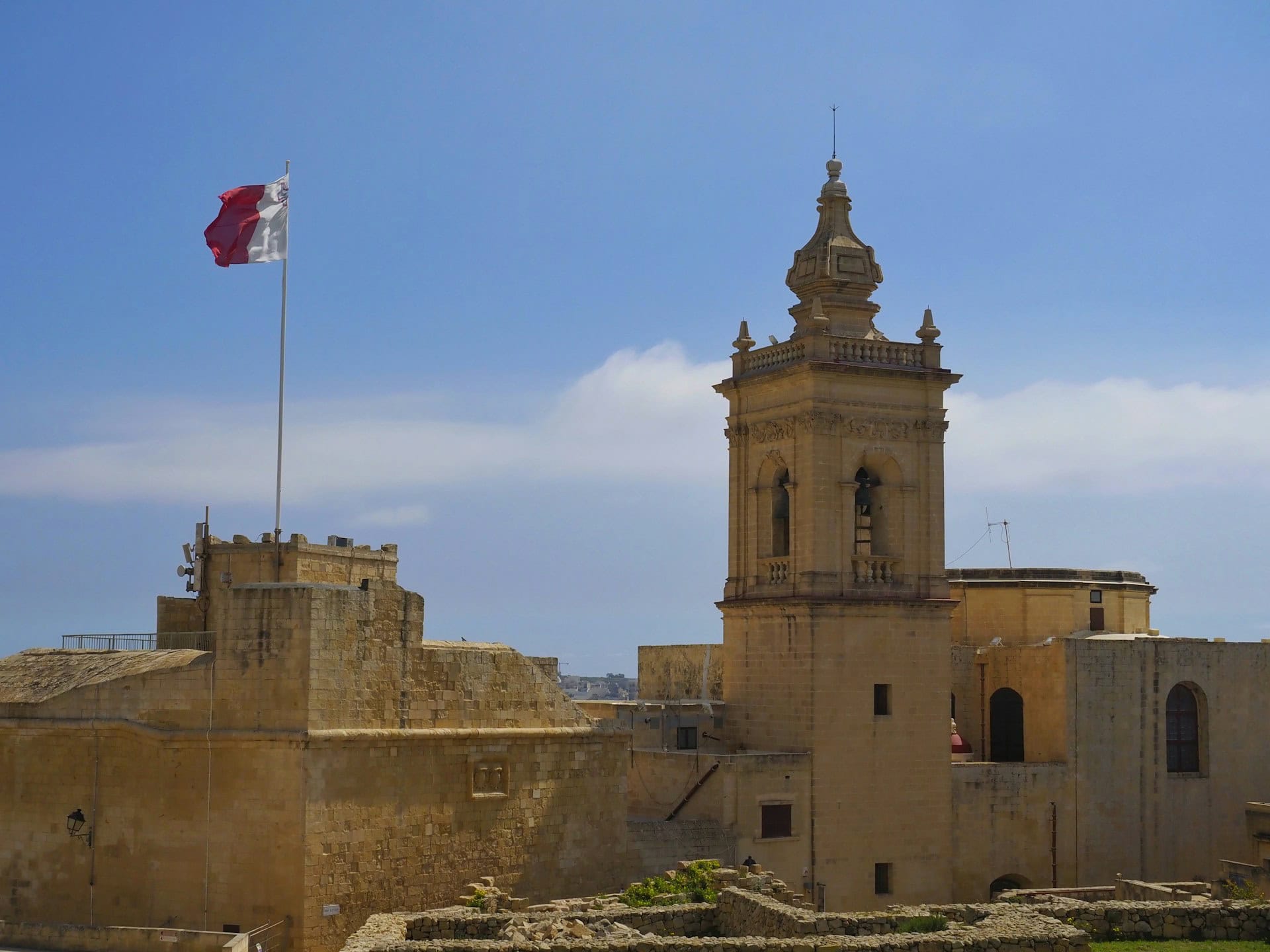 Blick auf die historische Cittadella in Victoria auf der maltesischen Insel Gozo. Eine große maltesische Flagge weht über den honigfarbenen Festungsmauern, daneben erhebt sich ein barocker Glockenturm aus Kalkstein. Im Vordergrund sind die alten Befestigungsanlagen zu sehen, darüber ein weiter Himmel mit dichten Wolken.