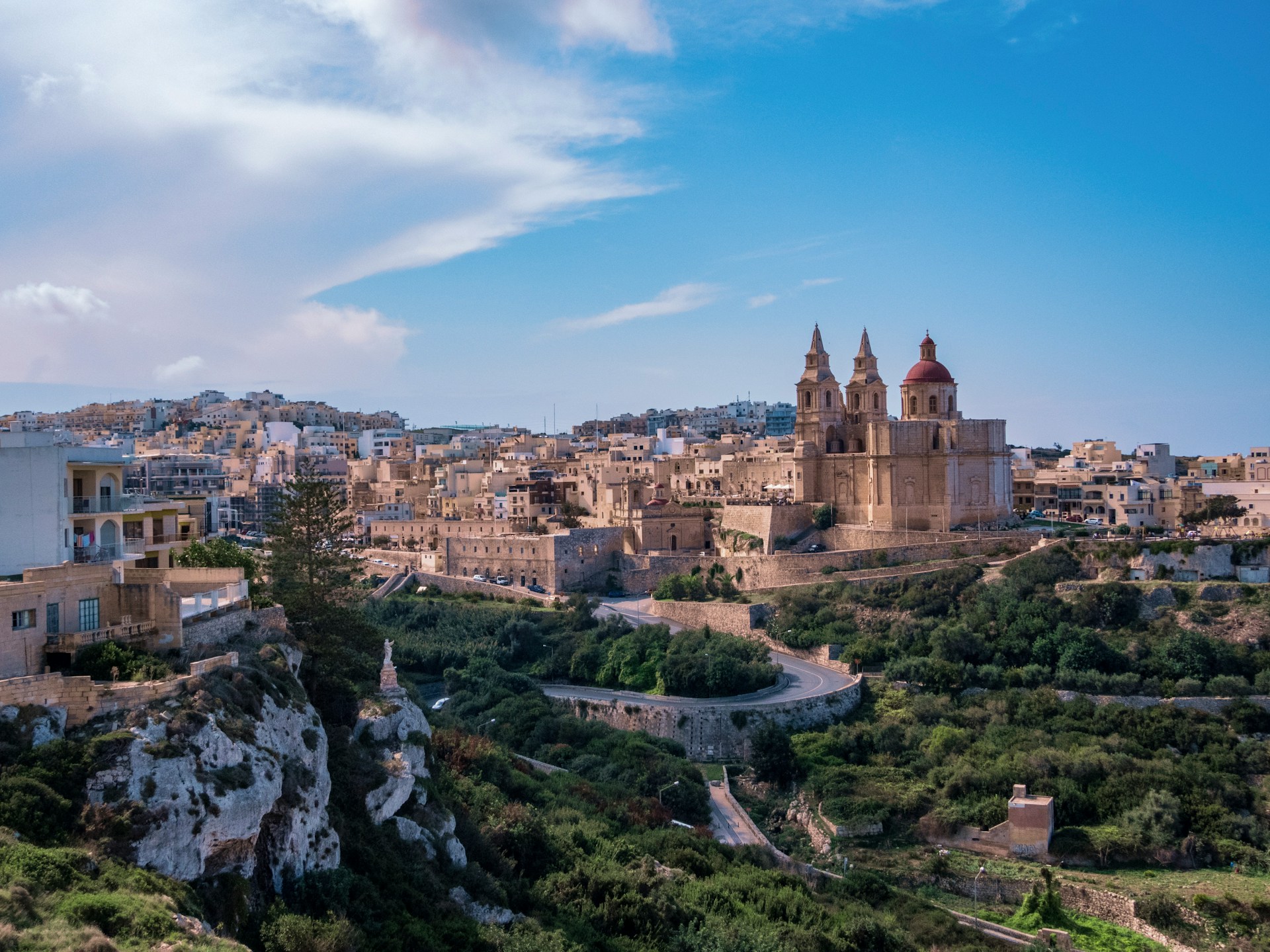 Panoramablick auf die historische Stadt Mdina auf Malta. Im Zentrum erhebt sich die barocke St.-Pauls-Kathedrale mit zwei Glockentürmen und einer roten Kuppel, umgeben von honigfarbenen Kalksteingebäuden und Stadtmauern. Im Vordergrund schlängelt sich eine Straße durch ein grünes Tal mit Felsen und Bäumen, darüber spannt sich ein weiter blauer Himmel mit dramatischen Wolken.