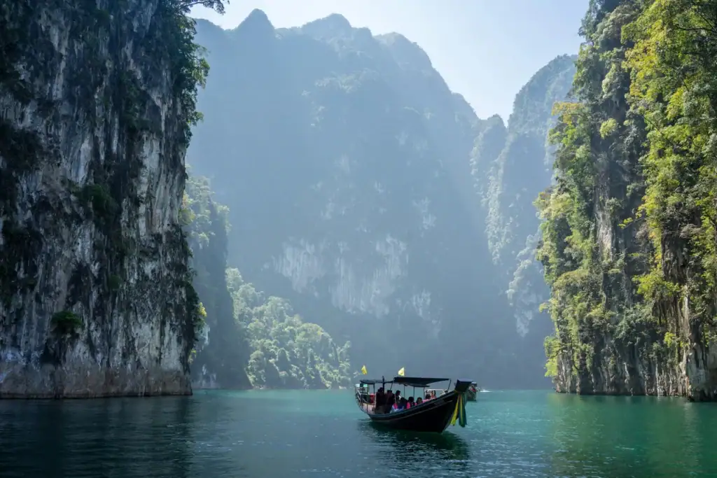 Ein traditionelles thailändisches Langschwanzboot mit Passagieren fährt auf dem smaragdgrünen Cheow-Lan-See im Khao-Sok-Nationalpark. Das Boot wird von steilen, dicht bewachsenen Kalksteinfelsen und im Dunst liegenden Bergen umrahmt.