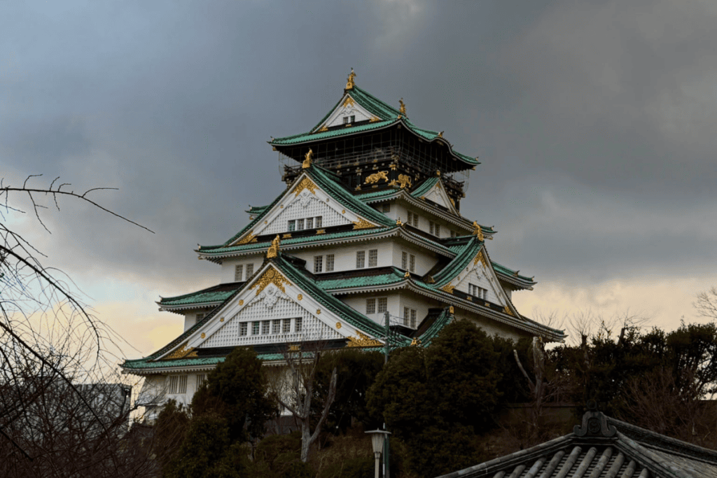 Nahaufnahme des mehrstöckigen Hauptturms der Burg Osaka mit grün-goldenen Dachverzierungen und weißen Wänden unter einem dramatischen, bewölkten Himmel.