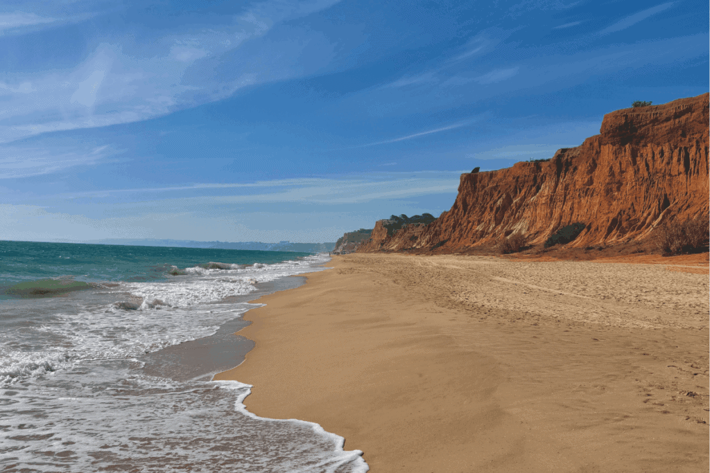 Blick entlang eines breiten, goldenen Sandstrandes mit auslaufendem weißem Wellenschaum auf der linken Seite und steil aufragenden, roterdenen Sandsteinfelsen auf der rechten Seite unter blauem Himmel.