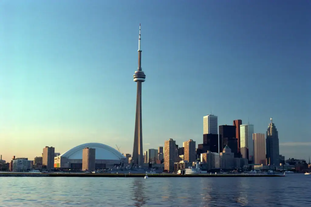 Skyline von Toronto bei klares Tageslicht mit dem markanten CN Tower im Zentrum, dem kuppelförmigen Rogers Centre Stadion auf der linken Seite und zahlreichen Wolkenkratzern vor blauem Himmel, gesehen über das Wasser des Ontario-Sees.