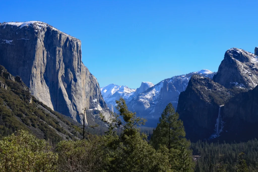 Granitwand El Capitan und schneebedeckte Gipfel über dem bewaldeten Yosemite Valley bei blauem Himmel