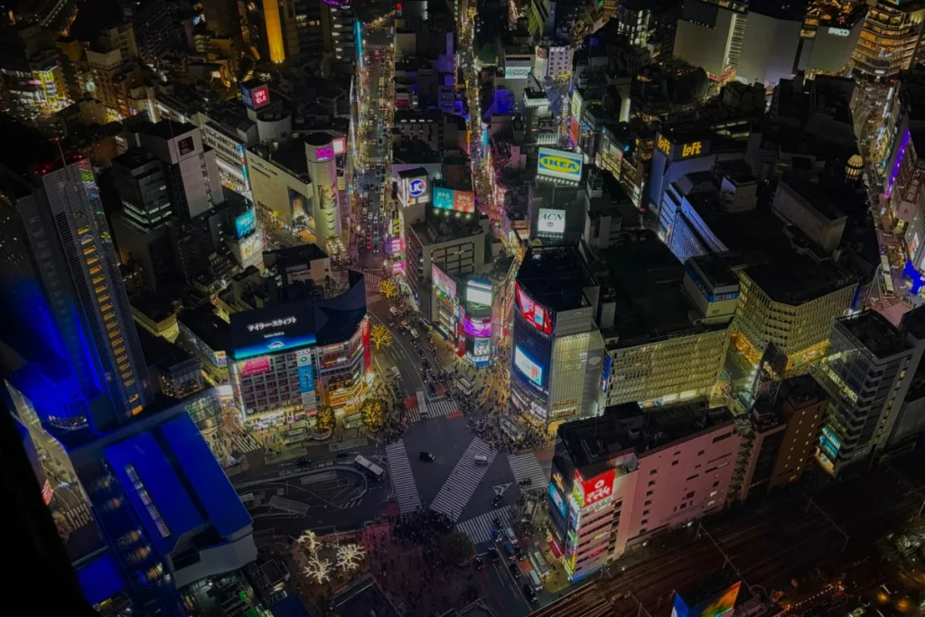 Nächtliche Luftaufnahme der beleuchteten Shibuya-Kreuzung in Tokio mit bunten Neonreklamen, Hochhäusern und Stadtverkehr aus der Vogelperspektive