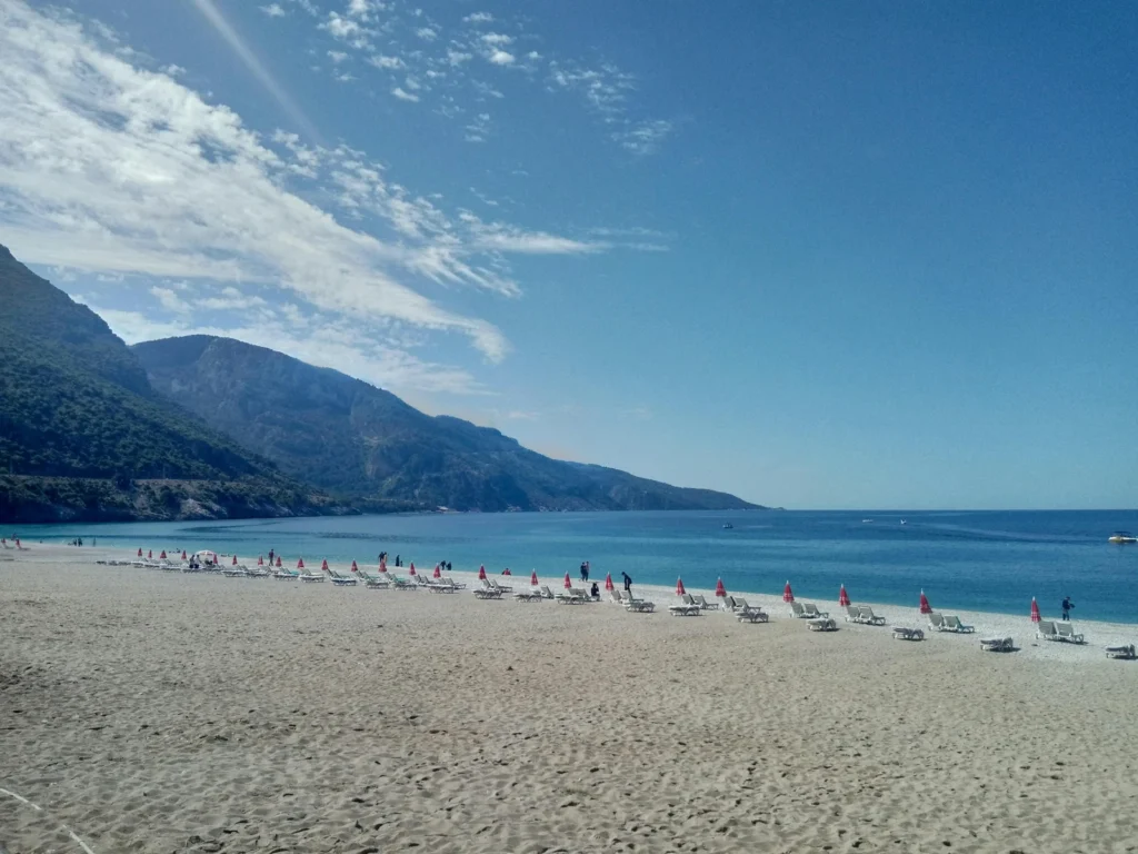 Langer Sandstrand von Ölüdeniz mit Liegen am türkisblauen Meer vor bewaldeten Bergen
